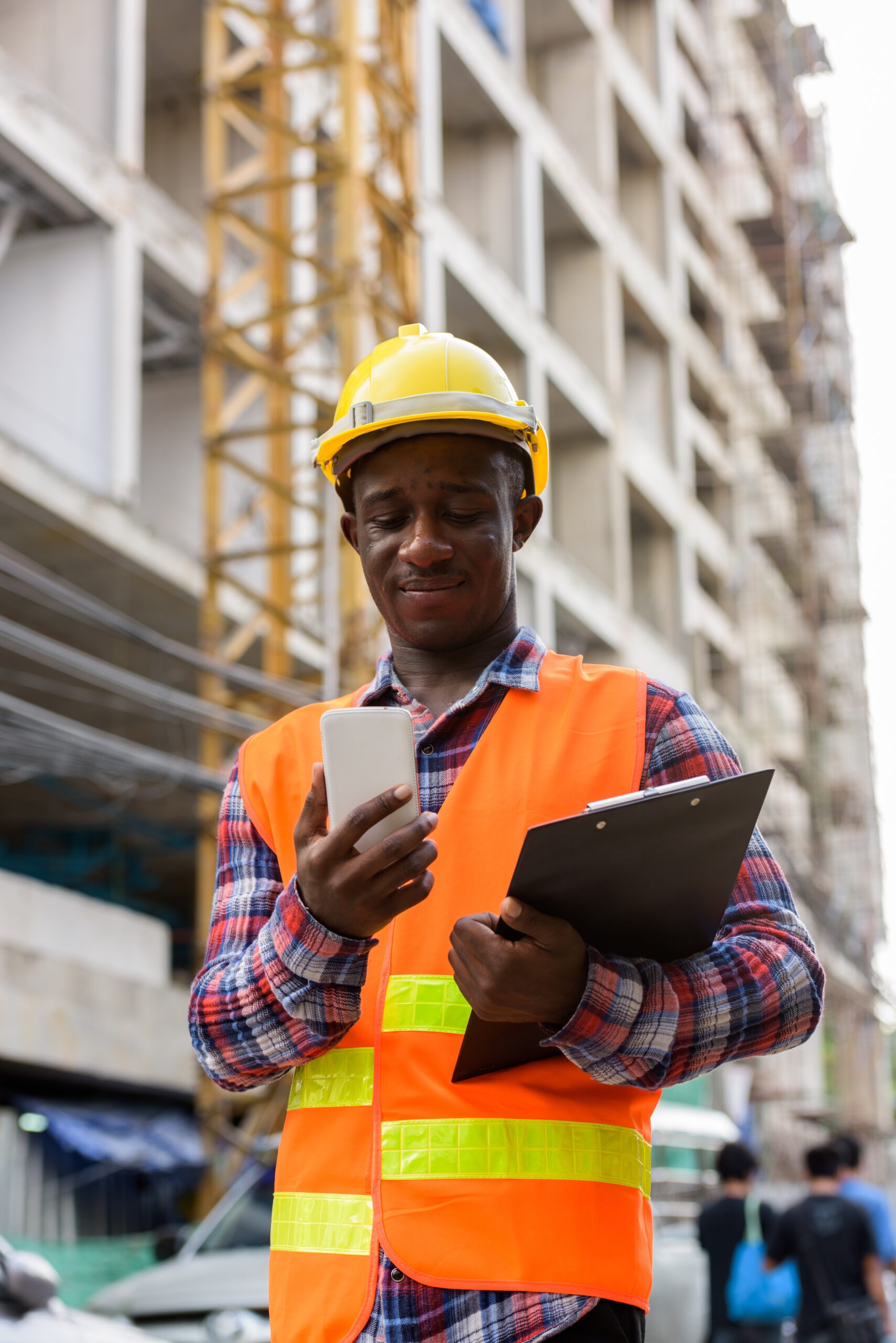 Young black African man construction worker holding clipboard while using mobile phone at building site vertical shot
