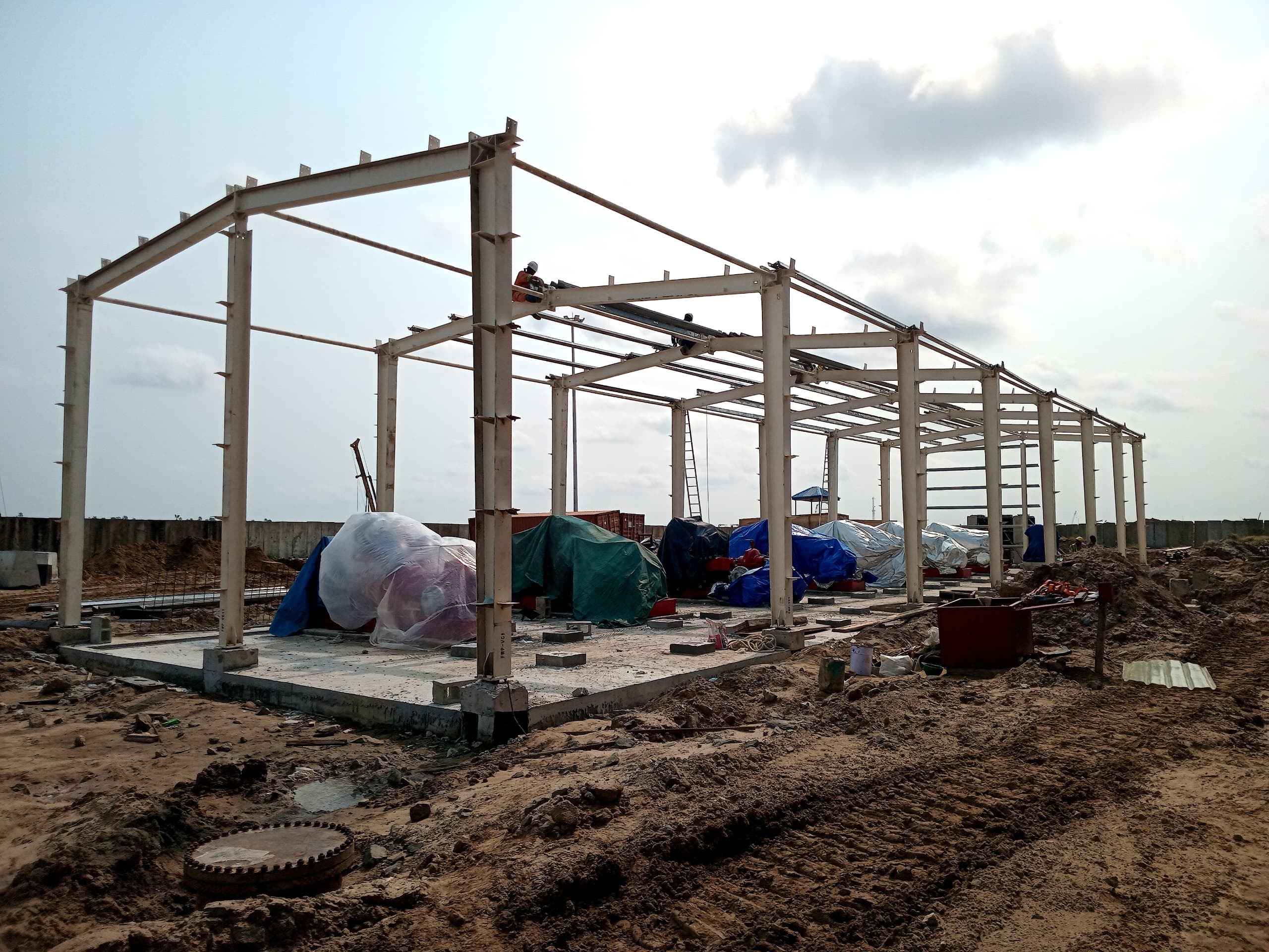 Steel framework of a partially constructed building on a construction site, surrounded by covered materials and equipment under a cloudy sky.