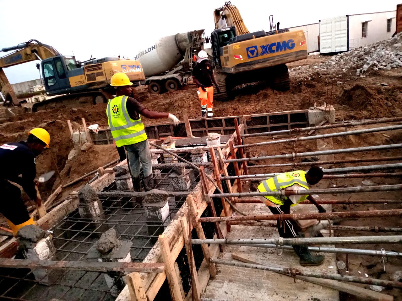 Construction workers in hard hats and reflective vests are pouring concrete into molds on a building site, with heavy machinery in the background.