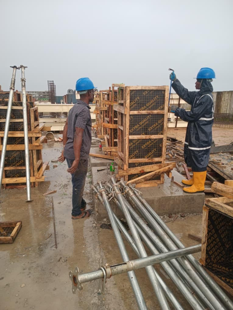 Two construction workers, one in a blue hard hat and the other in rain gear, inspect wooden formwork on a rainy site, surrounded by steel pipes.