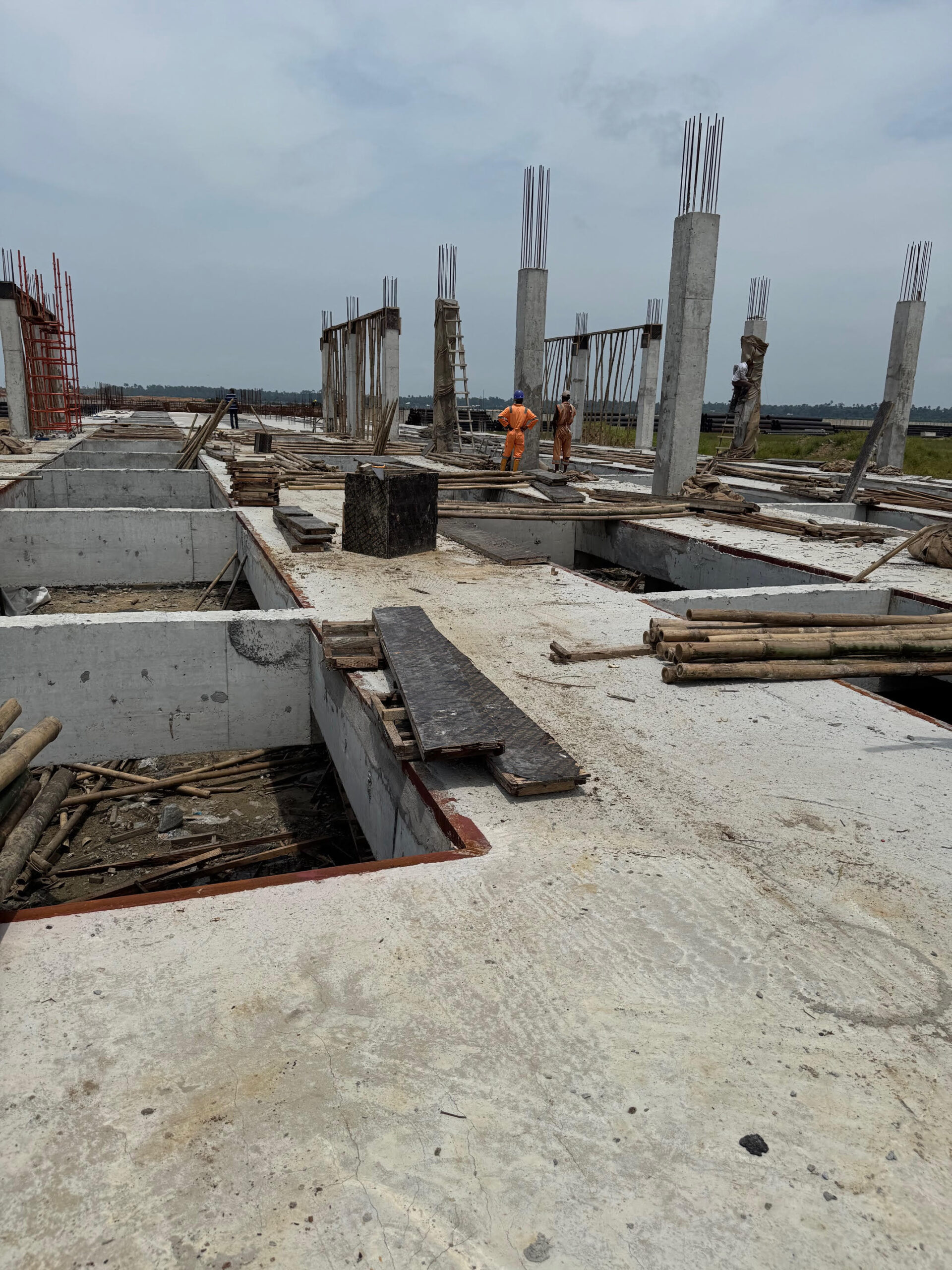Construction site featuring several concrete pillars and unfinished flooring, with workers in orange uniforms managing materials and scaffolding.