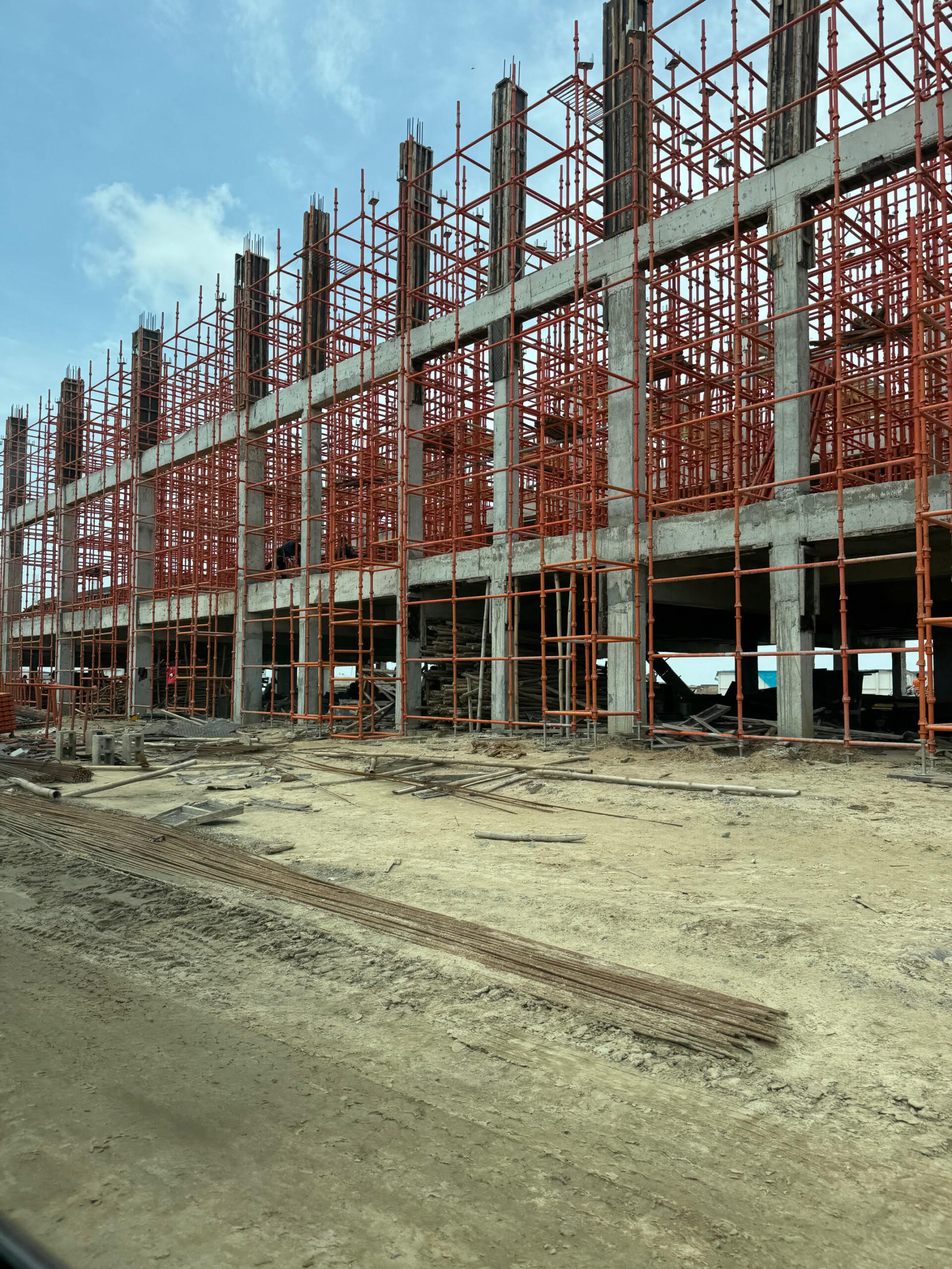 Construction site featuring a multi-story building framed with concrete pillars and covered in orange scaffolding, under a blue sky.