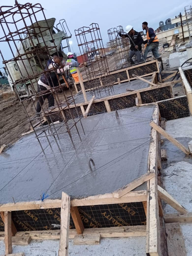 Construction workers are pouring concrete into wooden forms for a foundation, with rebar structures in the background, highlighting ongoing building activities.