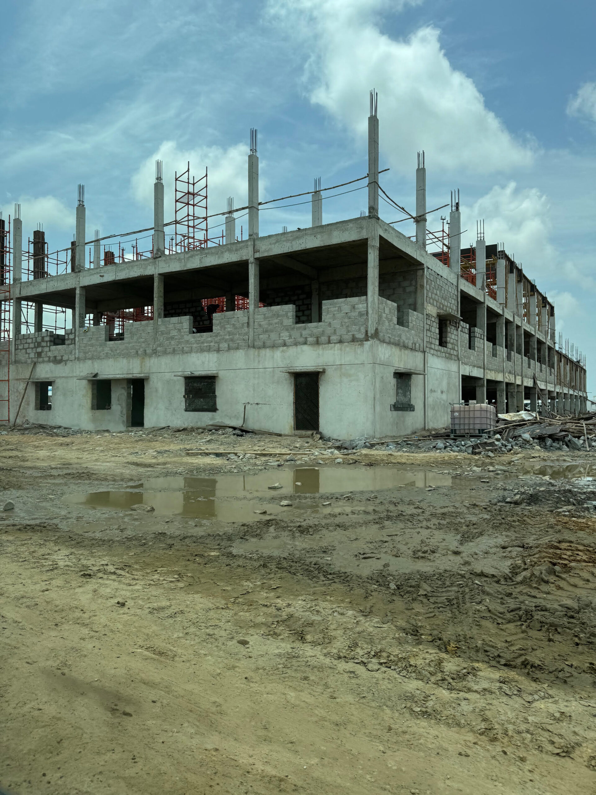Construction site featuring a partially built concrete structure with exposed columns and scaffolding against a cloudy sky. Puddles reflect the site’s progress.