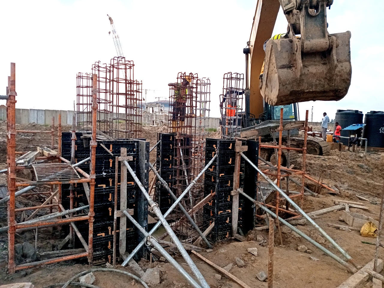 Construction site featuring rebar structures and formwork for concrete pouring, with an excavator lifting materials.