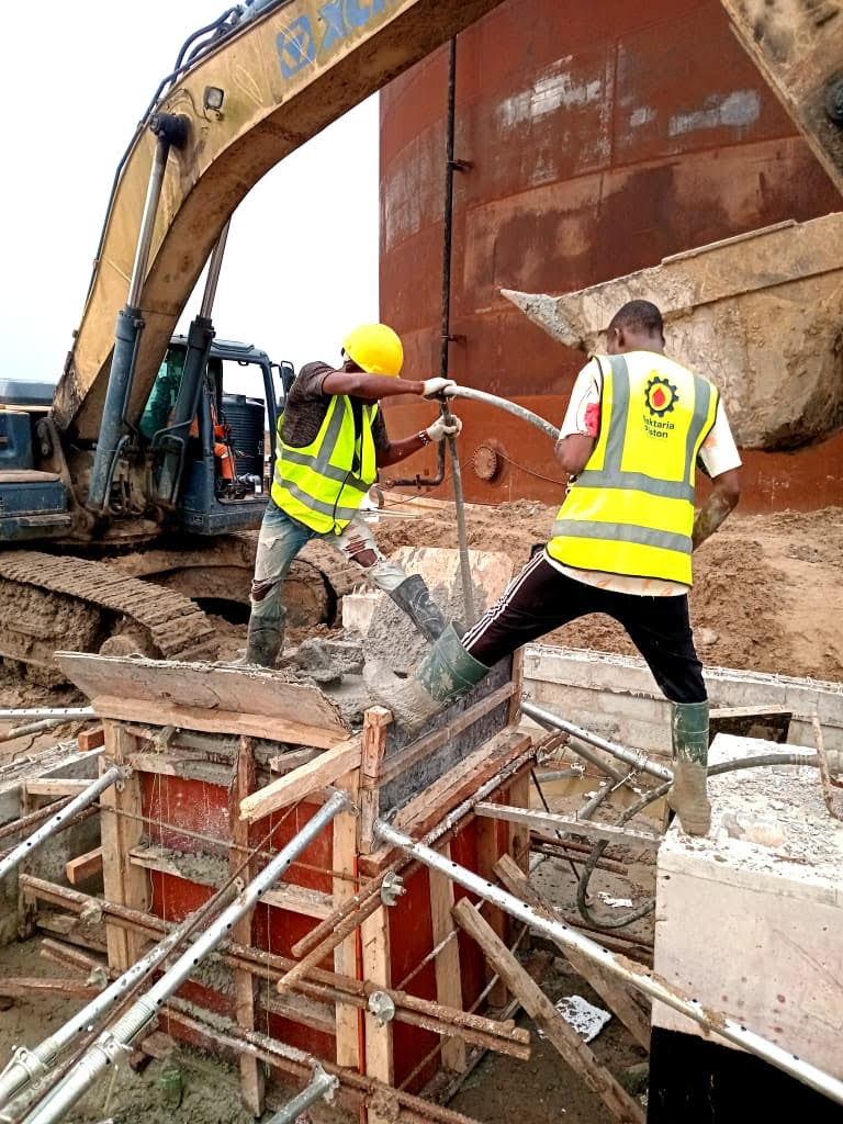 Construction workers in safety gear pour concrete into a wooden mold, with heavy machinery in the background, highlighting active site operations.