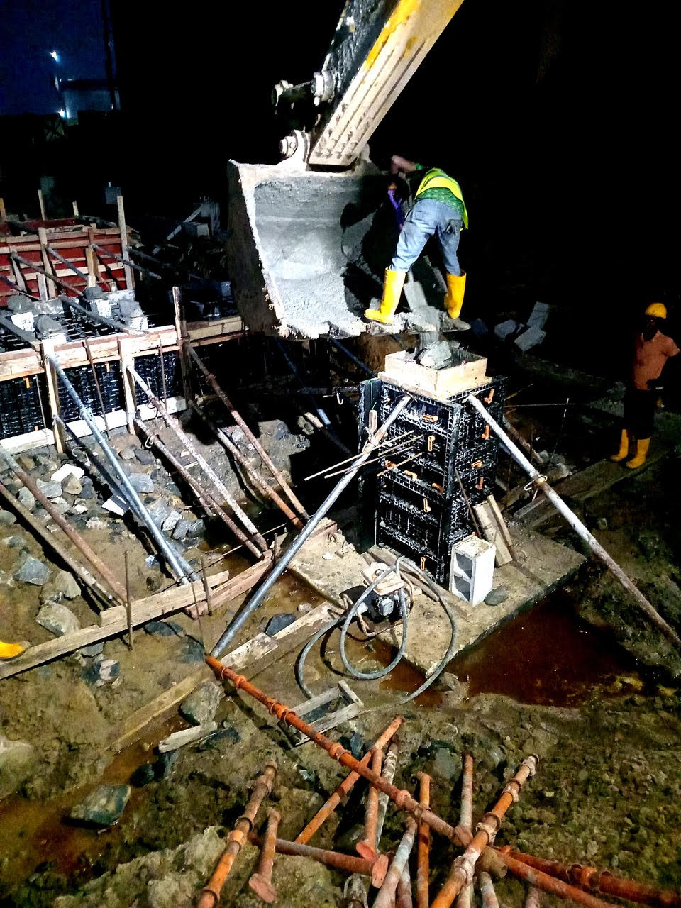 A construction worker uses a crane to pour concrete into a formwork at a nighttime construction site, surrounded by tools and materials.