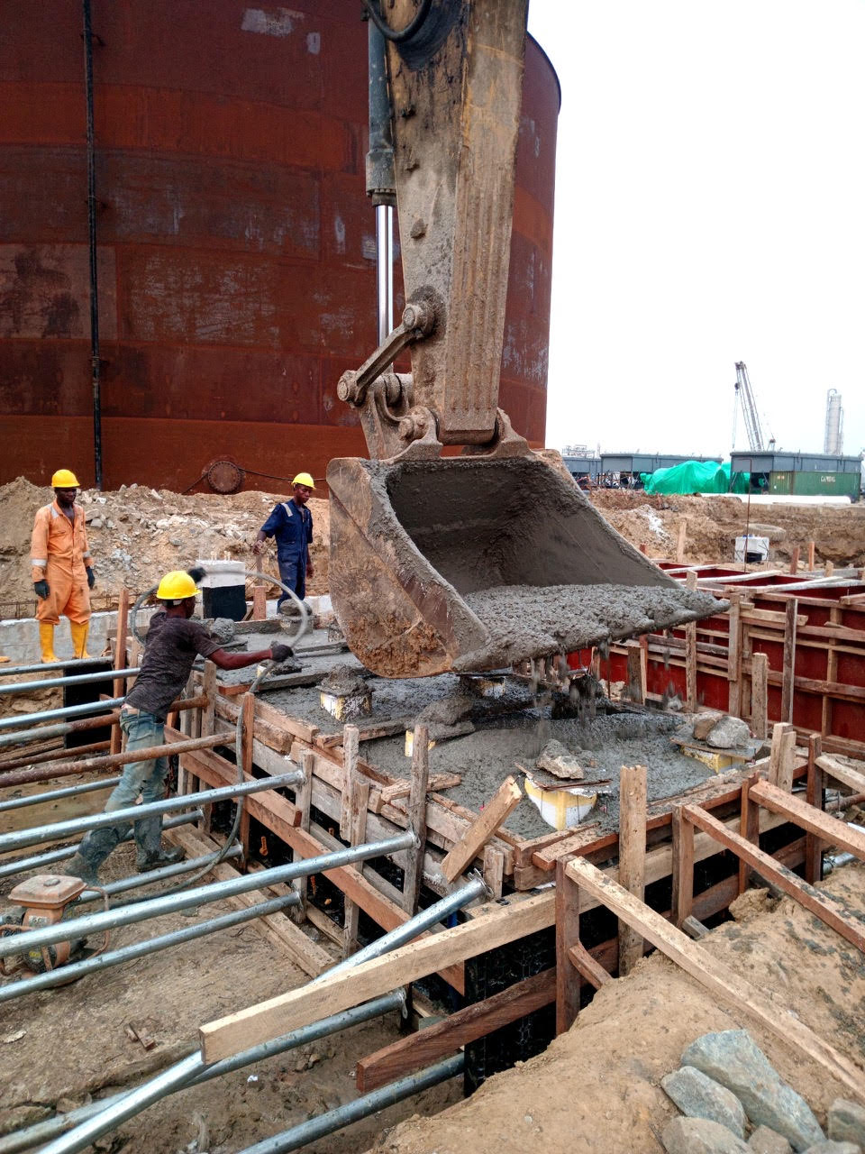 Construction workers pour concrete into wooden forms at a building site, with heavy machinery and a large storage tank in the background.