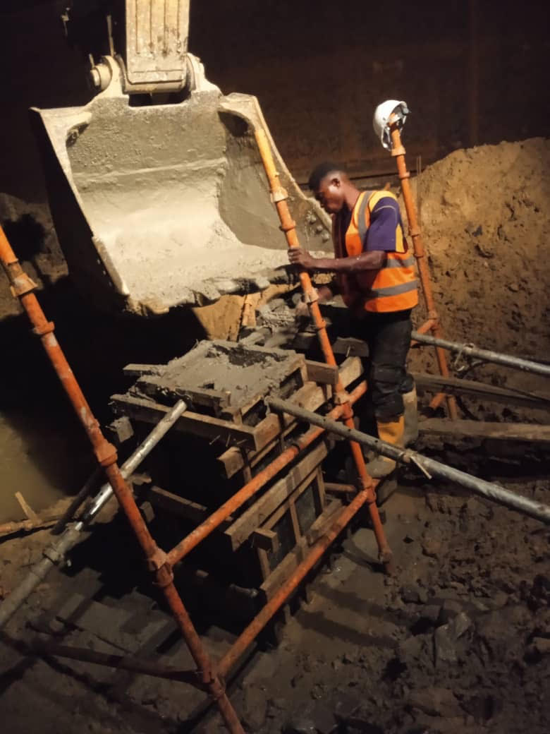 Construction worker in an underground site pours concrete into wooden molds, surrounded by earth and scaffolding for support.