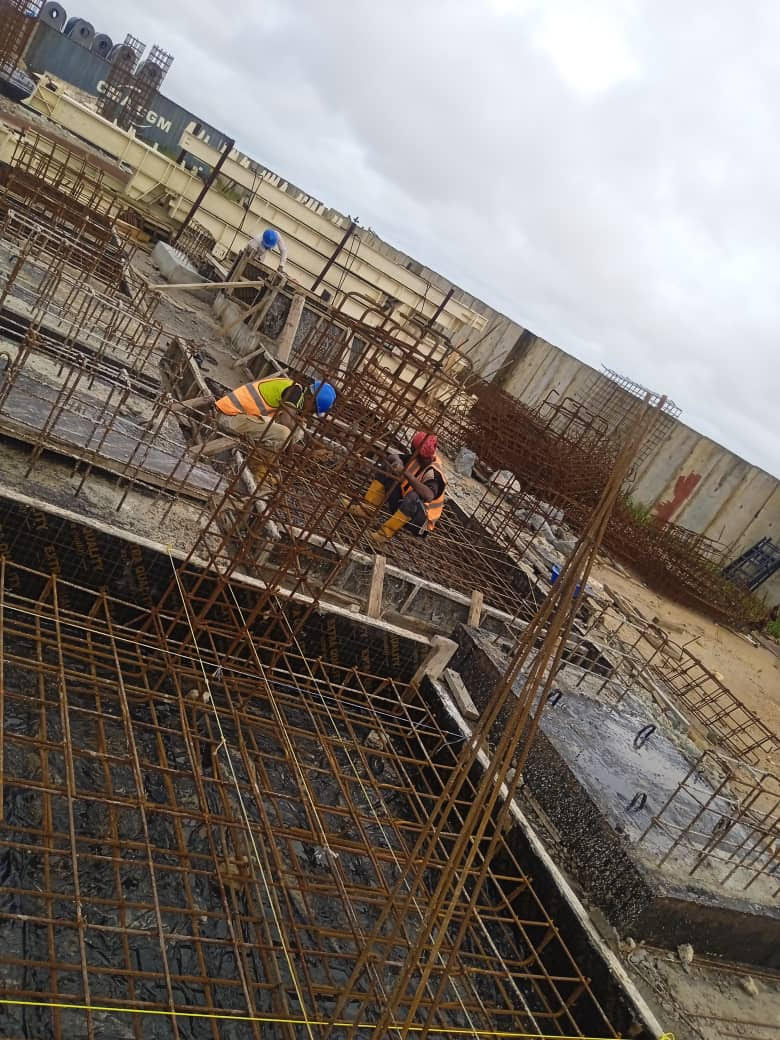 Construction workers install rebar in a foundation at a building site, with cloudy skies and construction materials visible in the background.