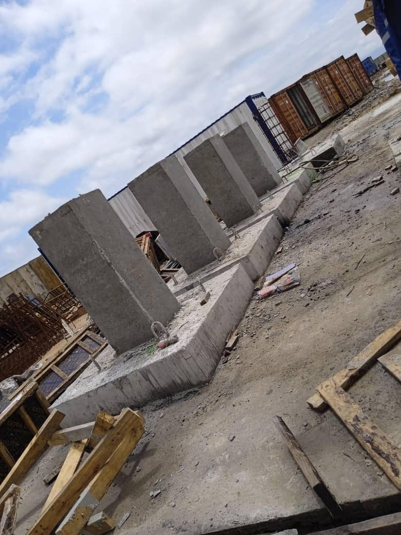 Concrete pillars stand on a construction site, partially embedded in a concrete base amidst scattered debris and storage containers.