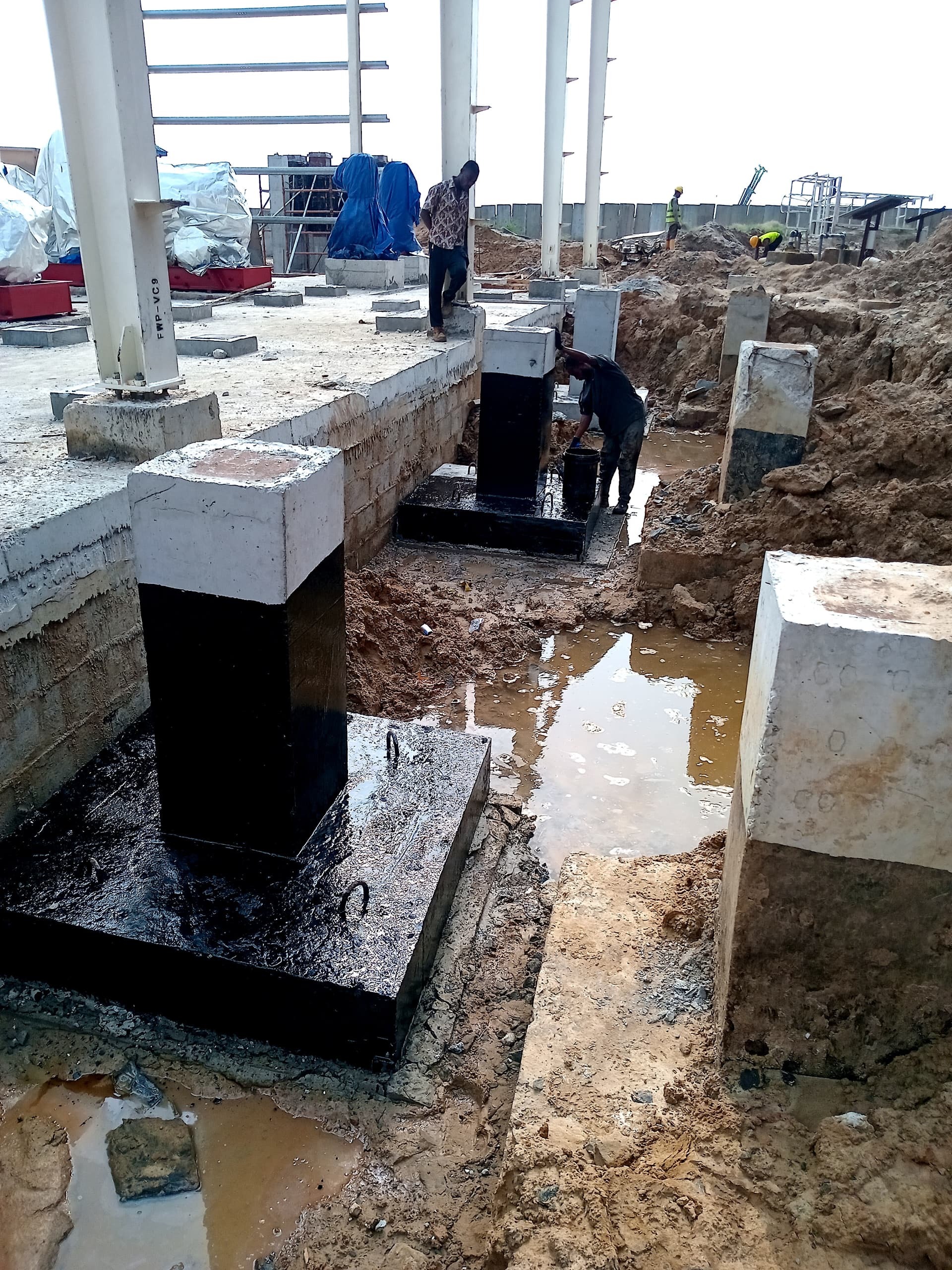 Construction site showing workers near concrete foundations with black bases in a muddy area, illustrating groundwork for a new structure.