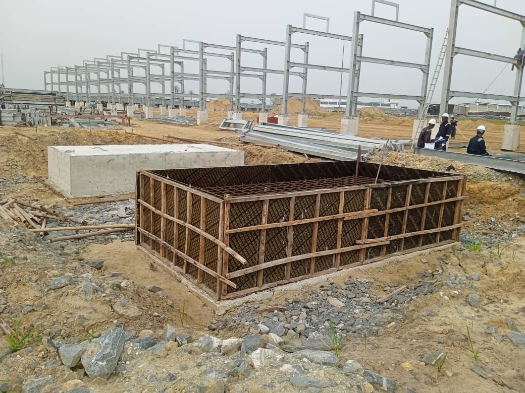 Construction site showing workers near concrete foundations with black bases in a muddy area, illustrating groundwork for a new structure.