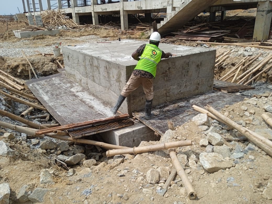 Construction worker in a hard hat and green vest secures a concrete foundation amidst a rocky, unfinished building site, emphasizing safety and diligence.
