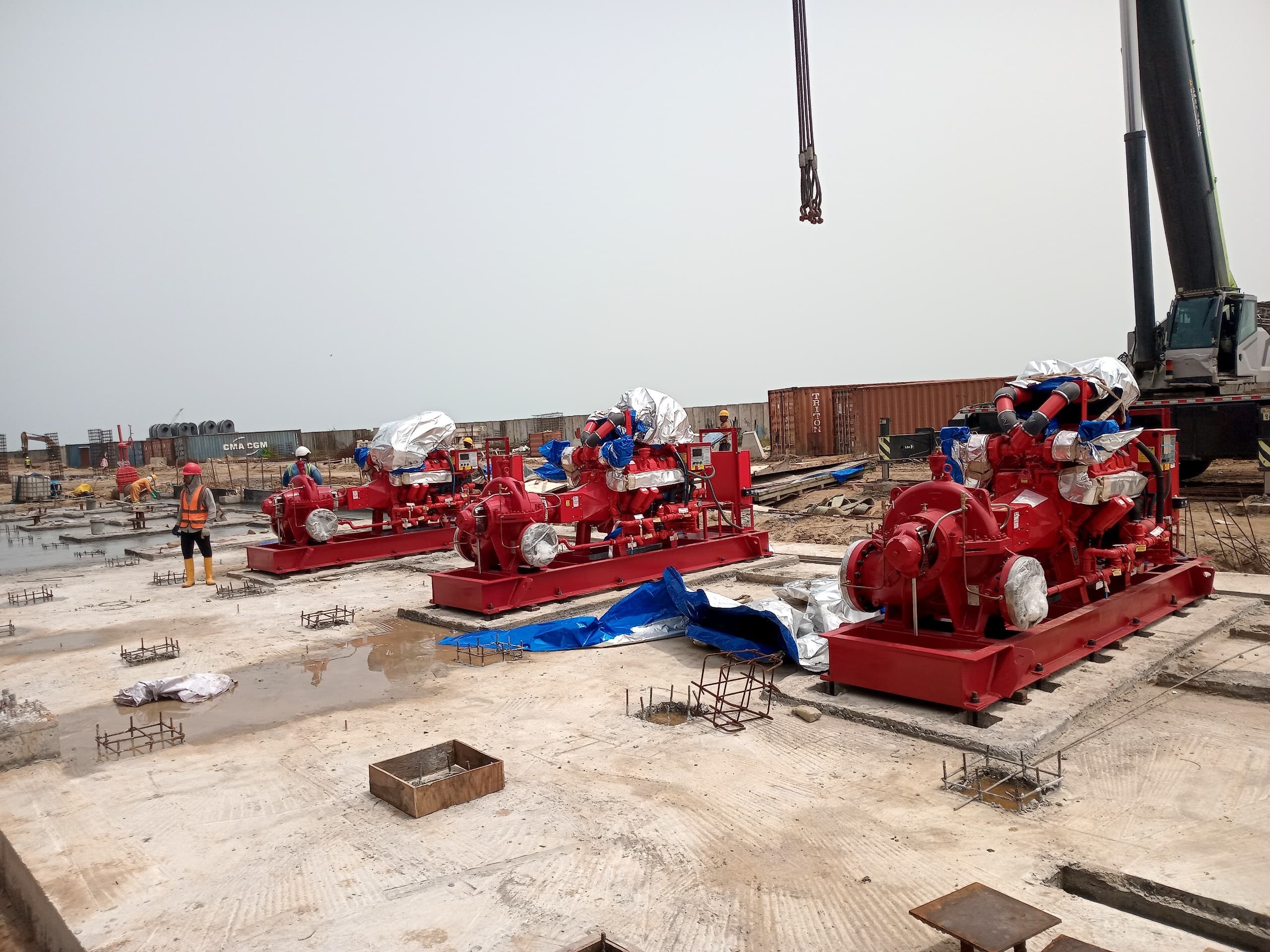 Three red industrial pumps are positioned on a construction site, surrounded by workers, materials, and machinery. A crane is visible above.